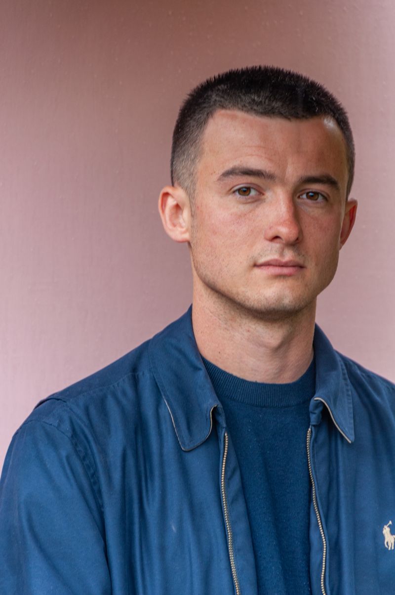 A person with closely cropped hair, in a blue zip jacket over a dark knit, facing the camera against a soft pink wall.