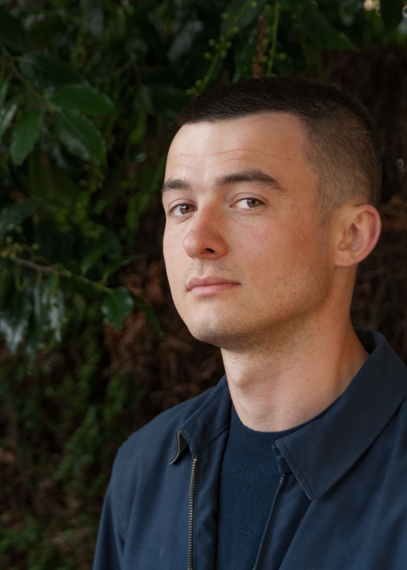 A person with cropped hair and a dark jacket turned slightly away, standing before dense green foliage in dappled shade.