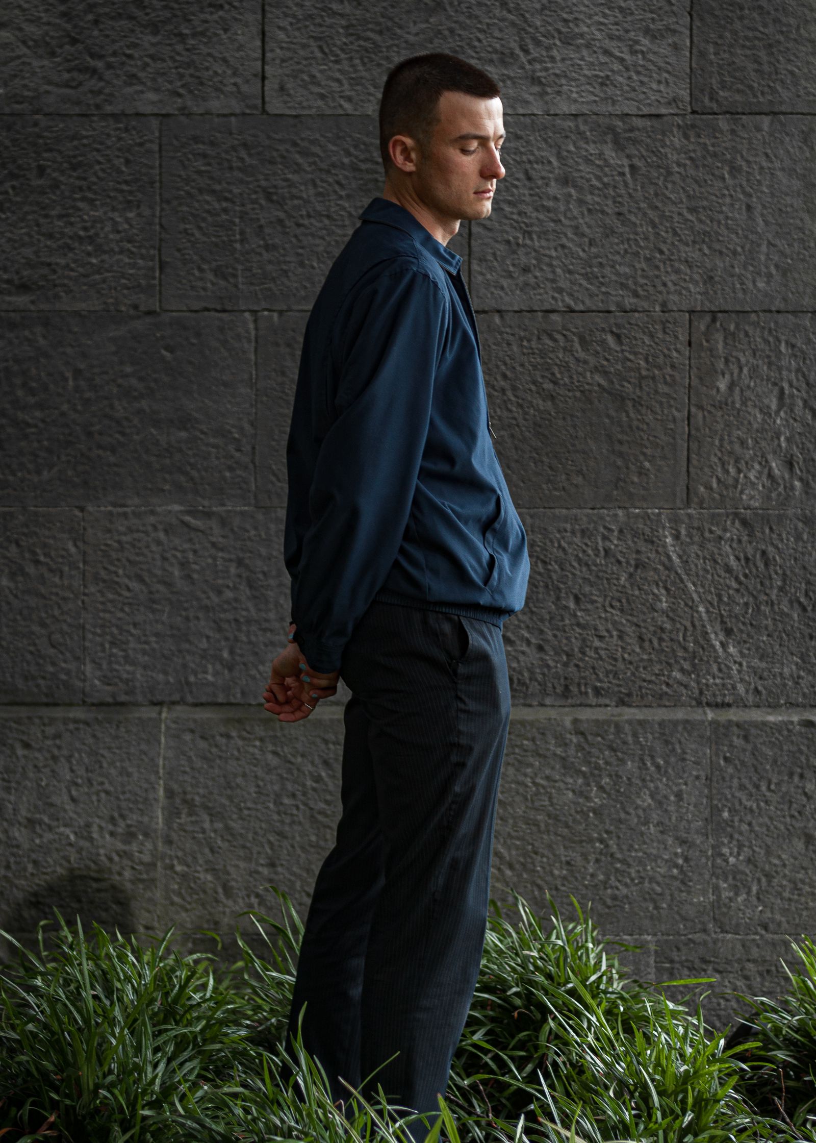 A person in a blue jacket and dark trousers standing in profile against a grey block wall, head lowered, hands clasped behind their back, grass at their feet.