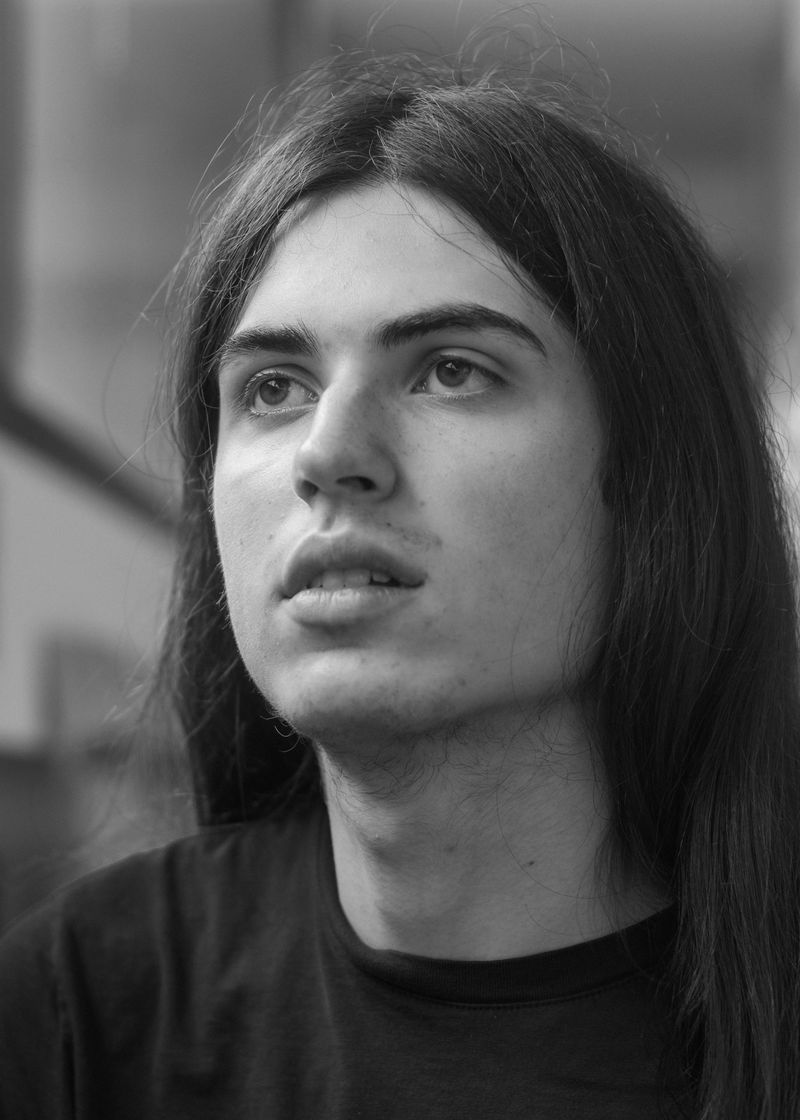 Black and white: a person with long hair and a dark shirt looking off to one side, railings blurred behind them.