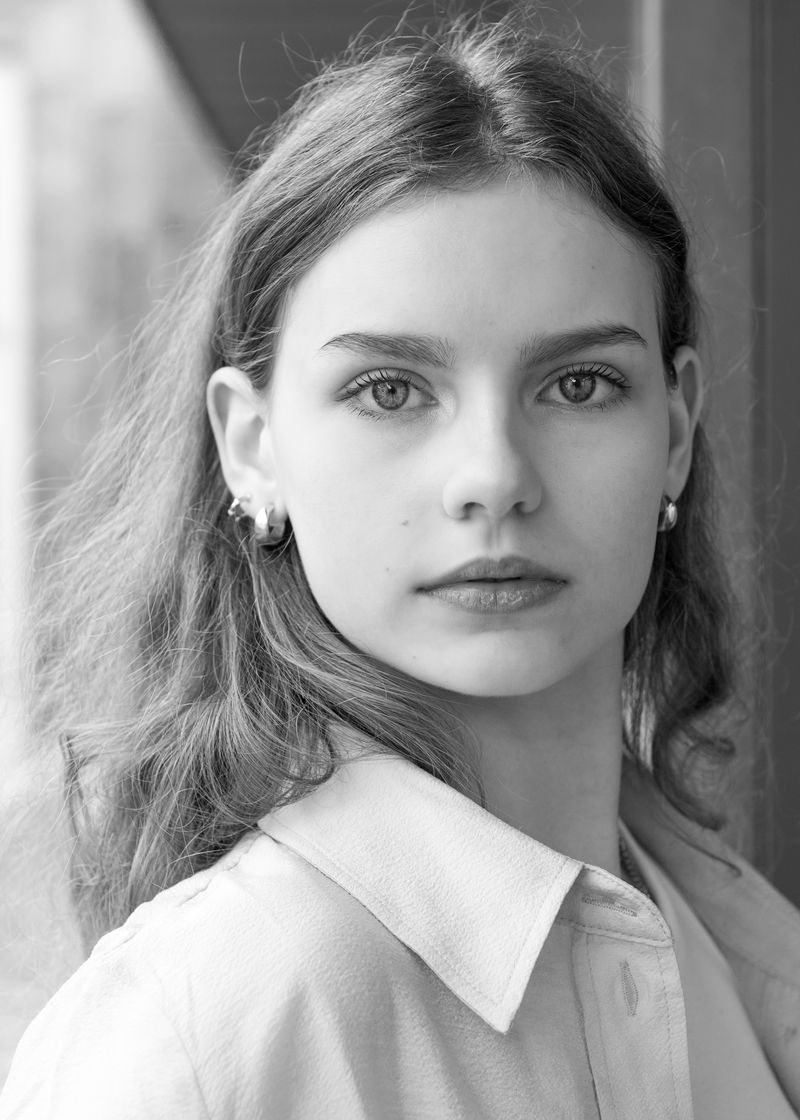 Black and white: a person with long hair and small drop earrings looking back over their shoulder, in a pale collared shirt.