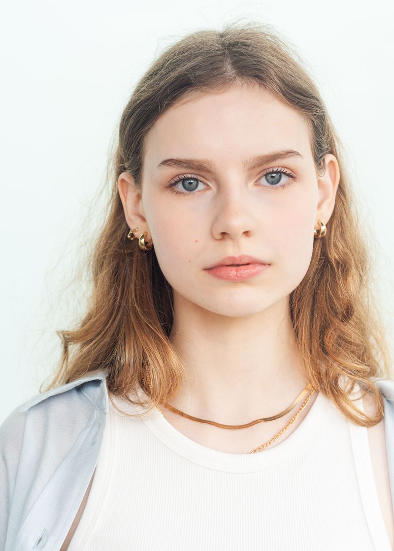 A person with long wavy hair, gold hoop earrings and a fine chain necklace, facing the camera in a white vest and pale jacket against a bright wall.