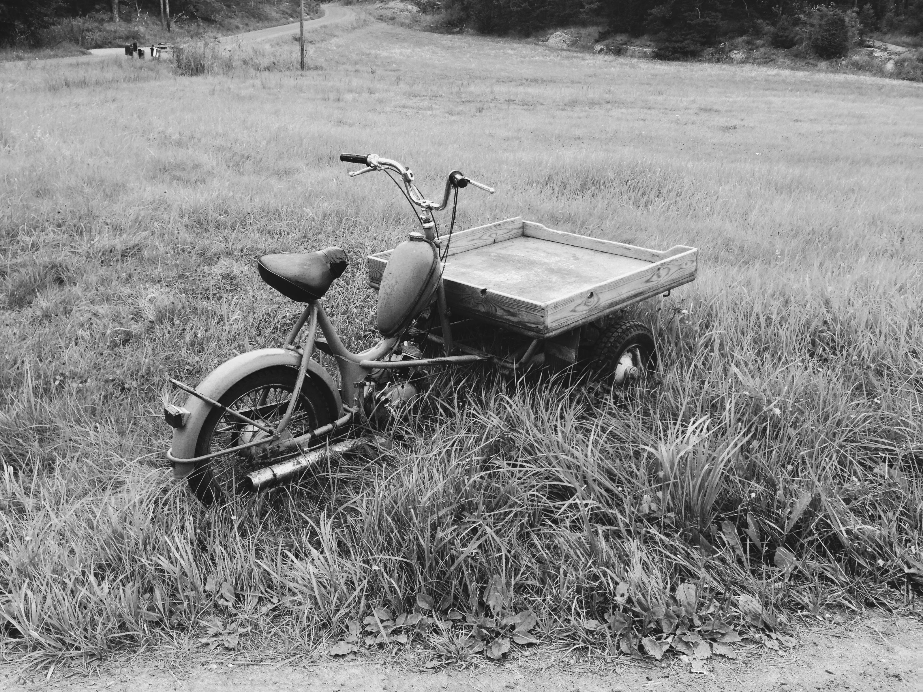Black and white: an old three-wheeled cargo moped with a wooden flatbed, parked in long grass beside a country road.