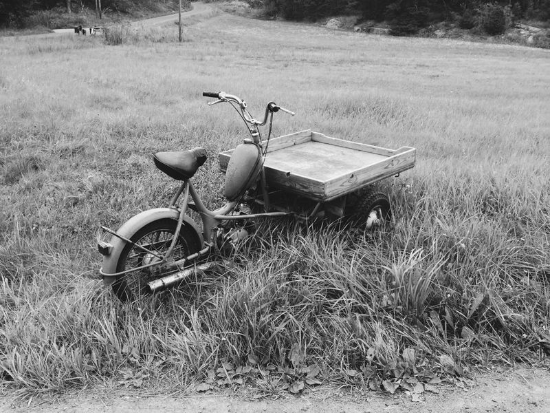 Black and white: an old three-wheeled cargo moped with a wooden flatbed, parked in long grass beside a country road.