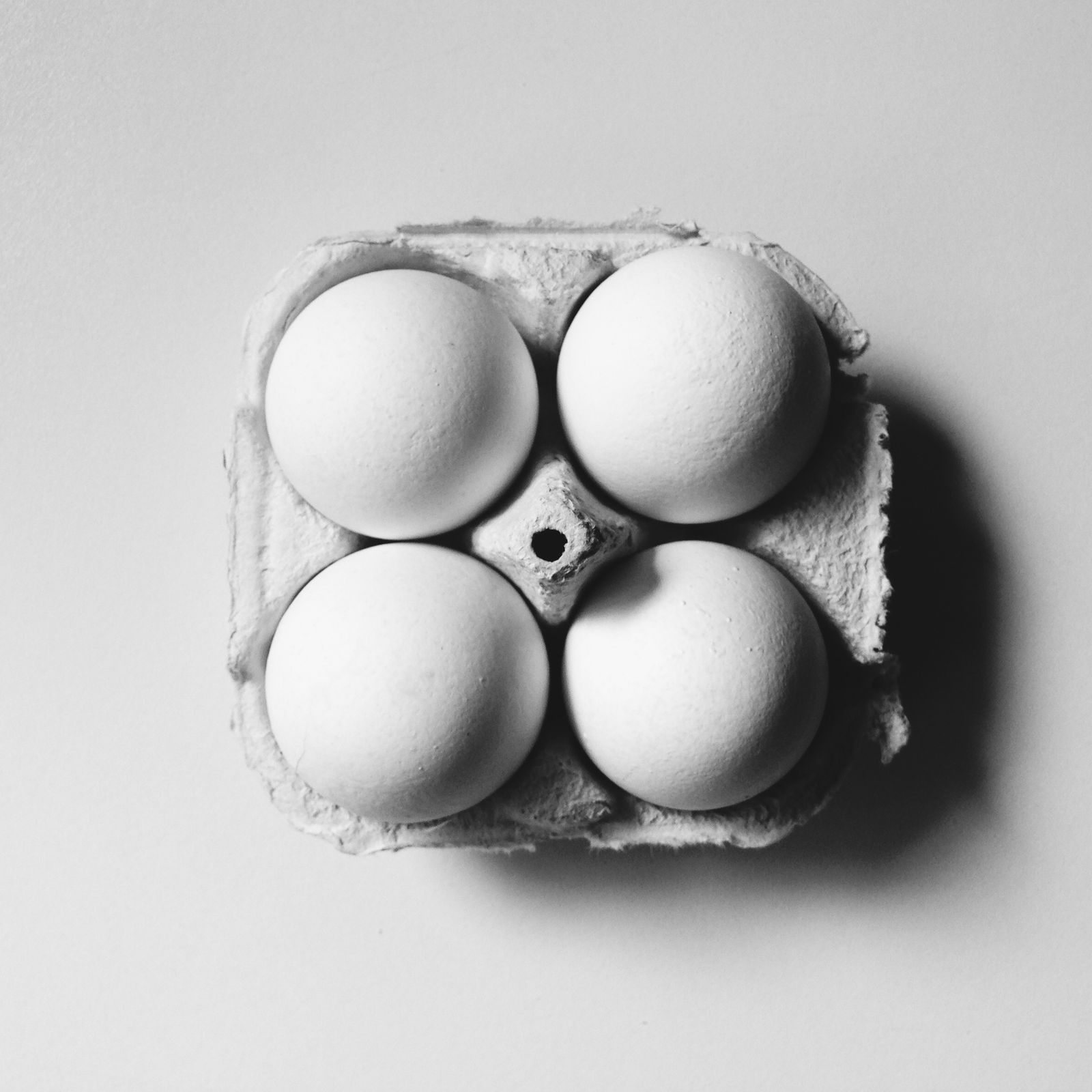 Black and white: four white eggs in a torn-open carton, seen from directly above on a pale surface.