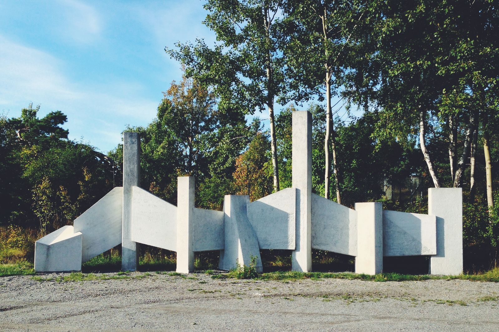 A white concrete sculpture of angled slabs and columns on gravel in front of birch trees, under a clear blue sky.