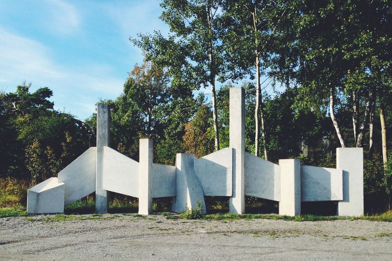 A white concrete sculpture of angled slabs and columns on gravel in front of birch trees, under a clear blue sky.
