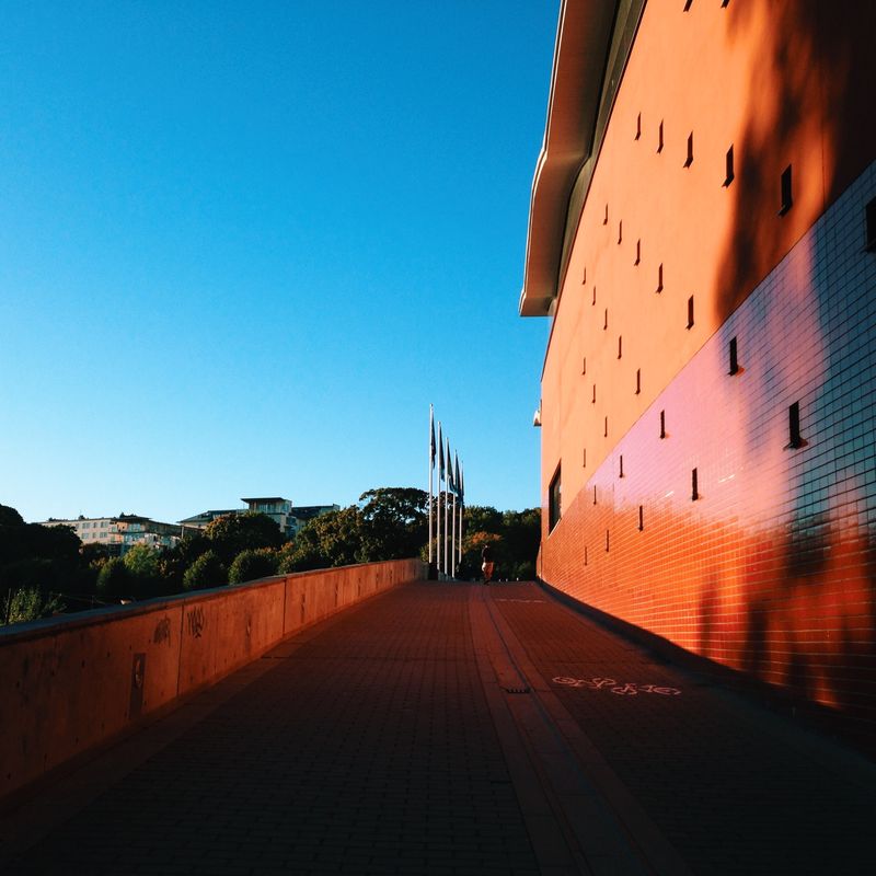 A brick-paved walkway rising alongside a building's orange-red tiled wall lit by low sun, flagpoles ahead under a clear blue sky.