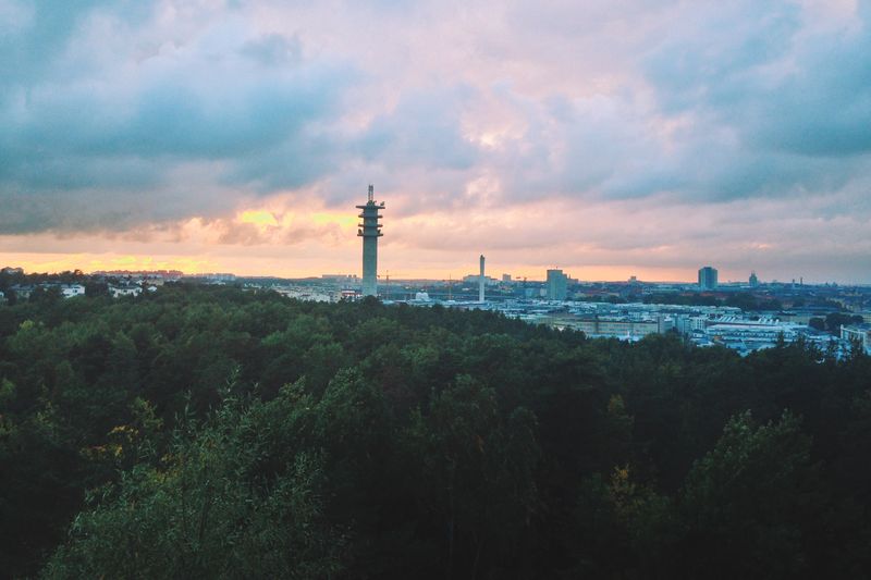 A city skyline at dusk under heavy clouds, a concrete telecom tower rising from dark forest, the horizon glowing orange.