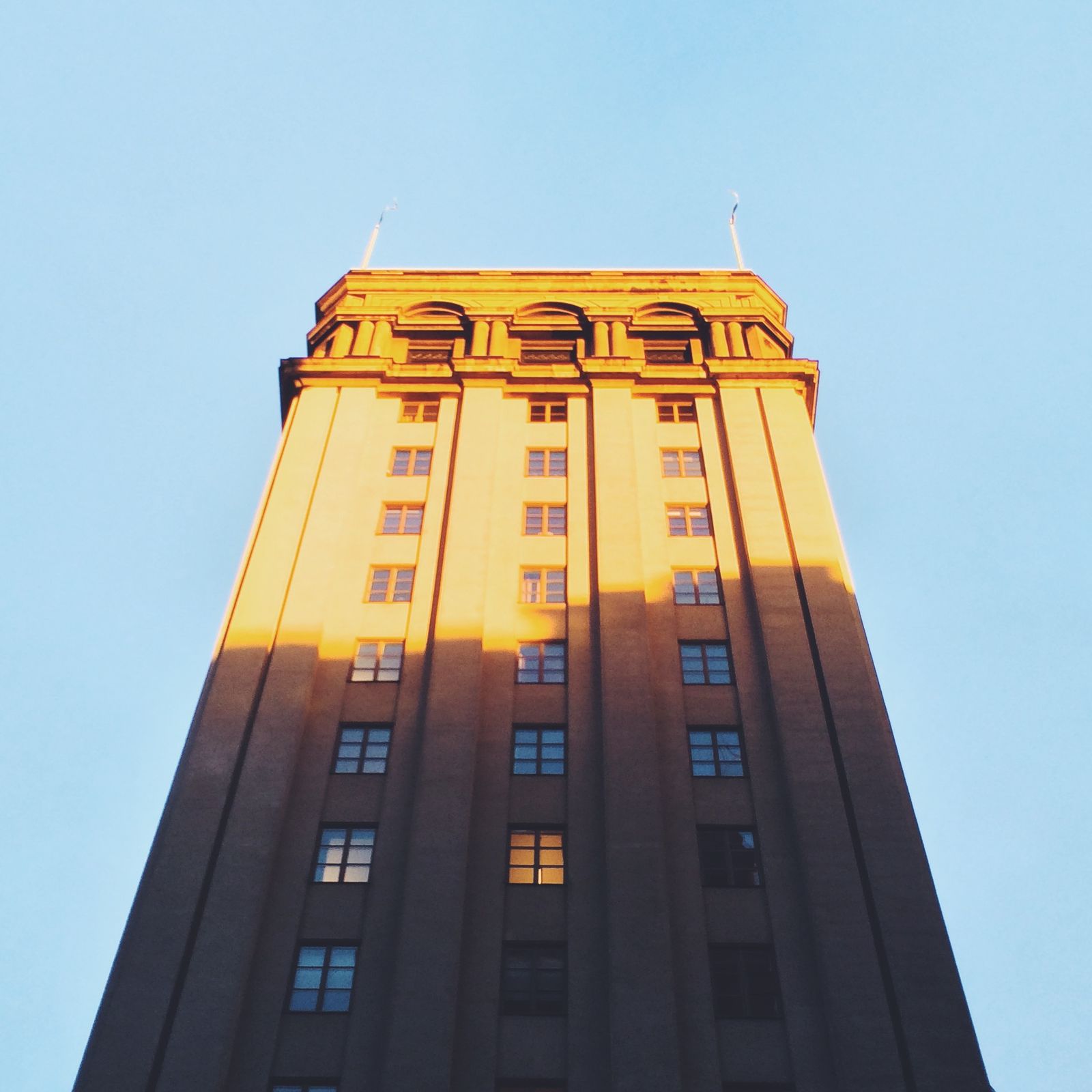 Looking straight up at a stone tower block, its arched crown lit gold by low sun against a clear sky, the lower floors in shadow.