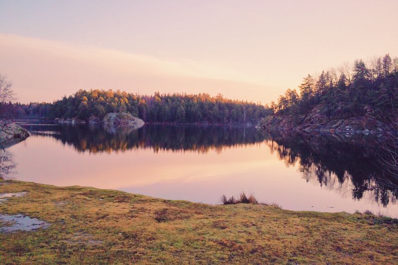 A still lake at dusk reflecting a pink sky, forested rocky shores on either side and frosty grass in the foreground.