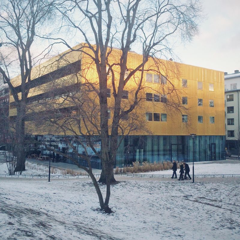 A gold-clad building with a glass ground floor beside a snowy park, bare trees in front and people walking past with a pram.