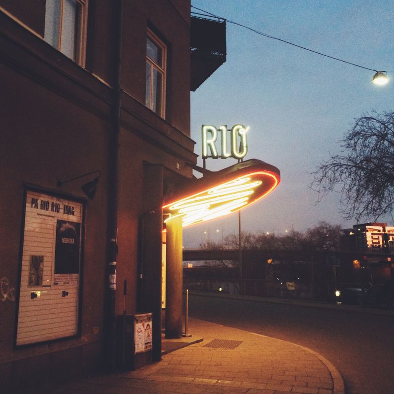 A cinema entrance at dusk, its neon sign and swept canopy glowing warm against a deep blue sky, a film listing board on the wall.