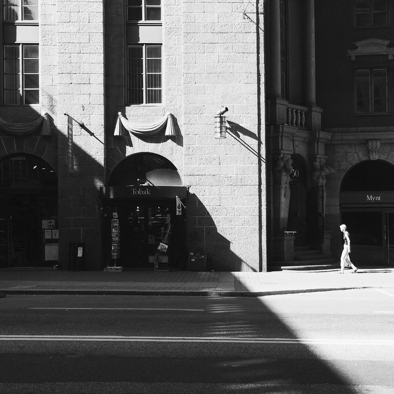 Black and white: a grand stone facade with arched shopfronts, a lone pedestrian crossing a sharp diagonal band of sunlight.