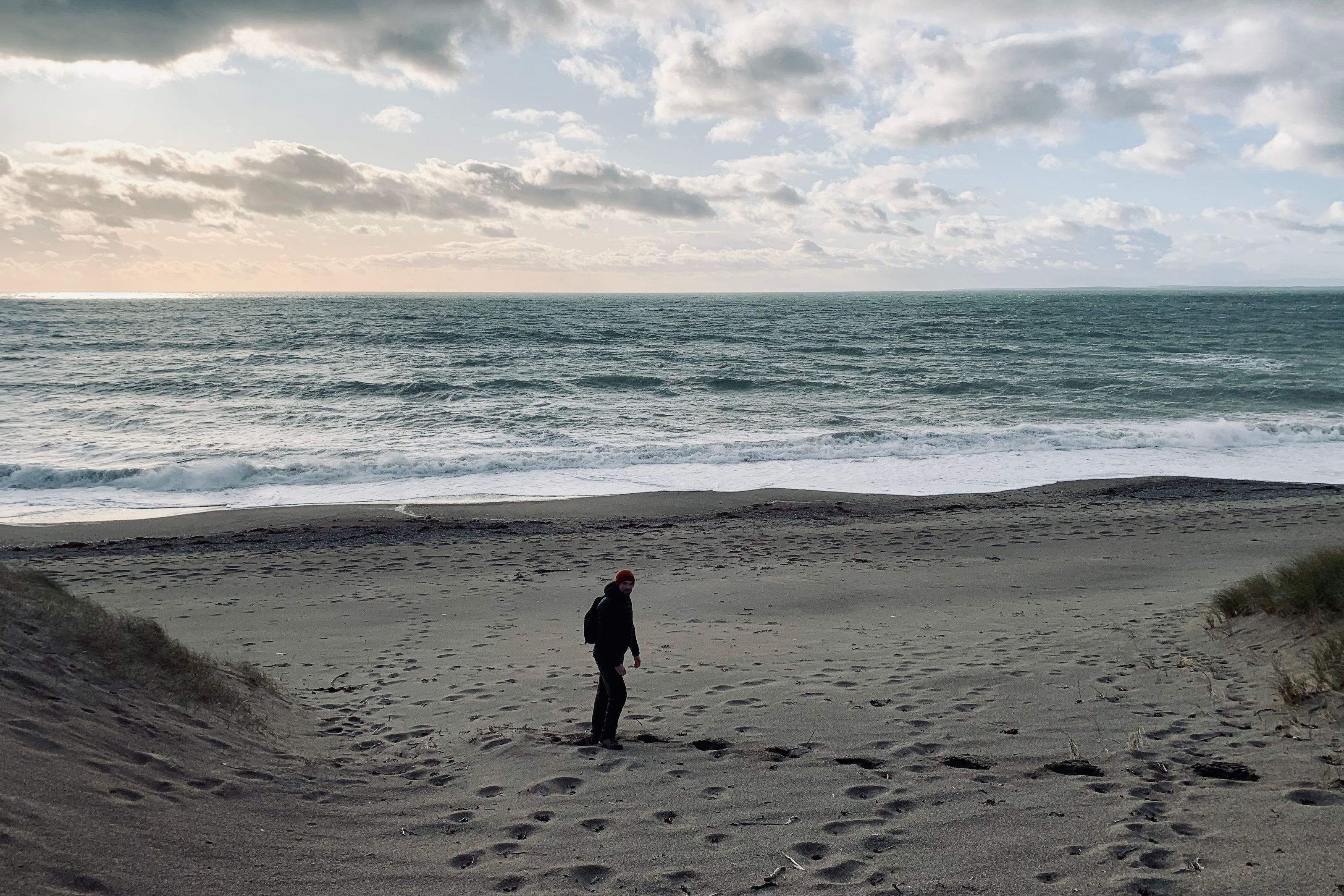 A figure in a beanie hat walking down a dune path onto a beach, rough grey surf and a cloud-streaked sky beyond.