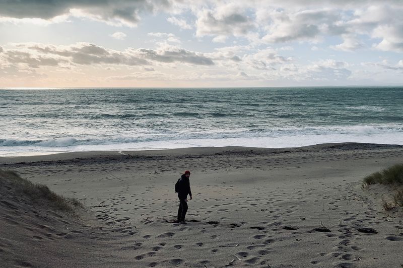 A figure in a beanie hat walking down a dune path onto a beach, rough grey surf and a cloud-streaked sky beyond.