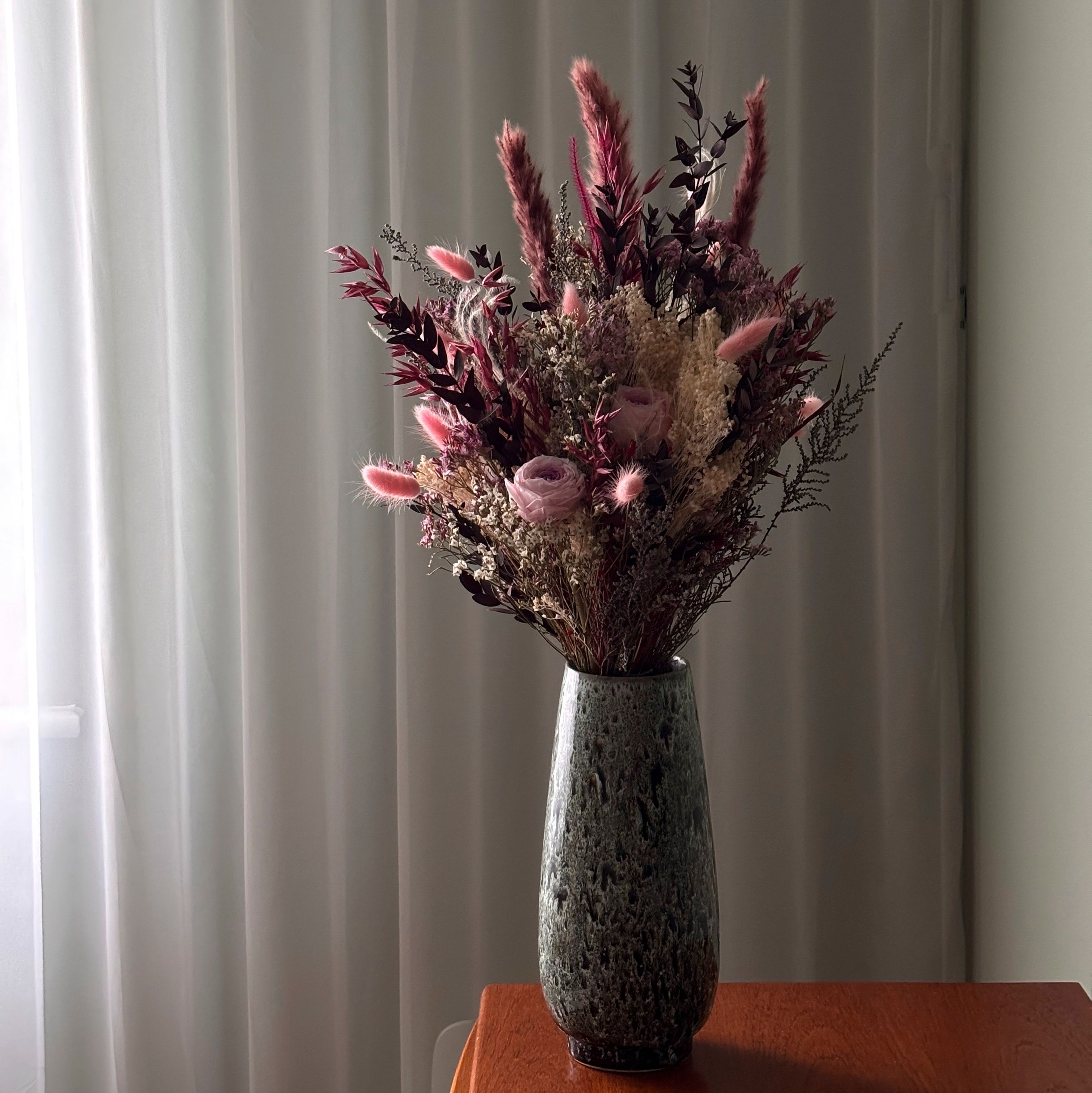A tall vase of dried flowers — pink roses, bunny tails and dark seed heads — standing on a wooden sideboard against a sunlit net curtain.