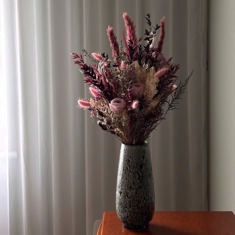A tall vase of dried flowers — pink roses, bunny tails and dark seed heads — standing on a wooden sideboard against a sunlit net curtain.