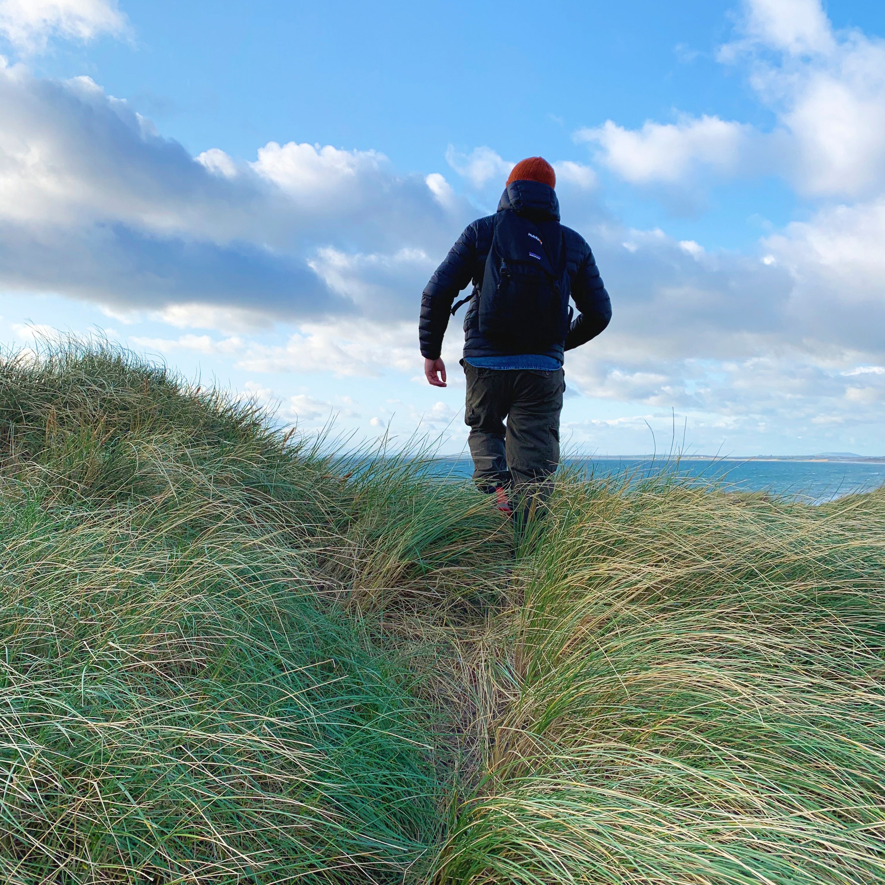 A person in a dark jacket and orange hat walking away through marram grass on a dune top, the sea and a bright, cloudy sky beyond.