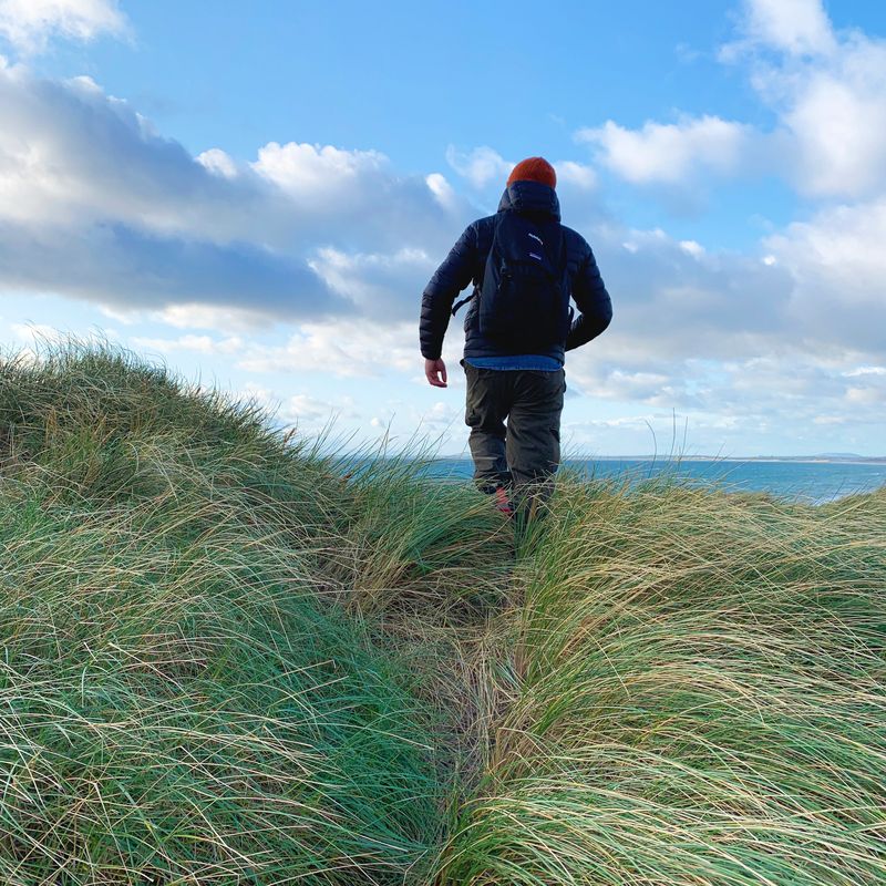 A person in a dark jacket and orange hat walking away through marram grass on a dune top, the sea and a bright, cloudy sky beyond.