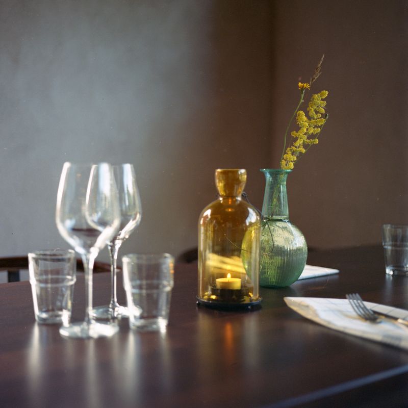 A restaurant table laid with wine glasses and tumblers, a candle burning inside an amber bottle, and a sprig of yellow flowers in a green glass vase.