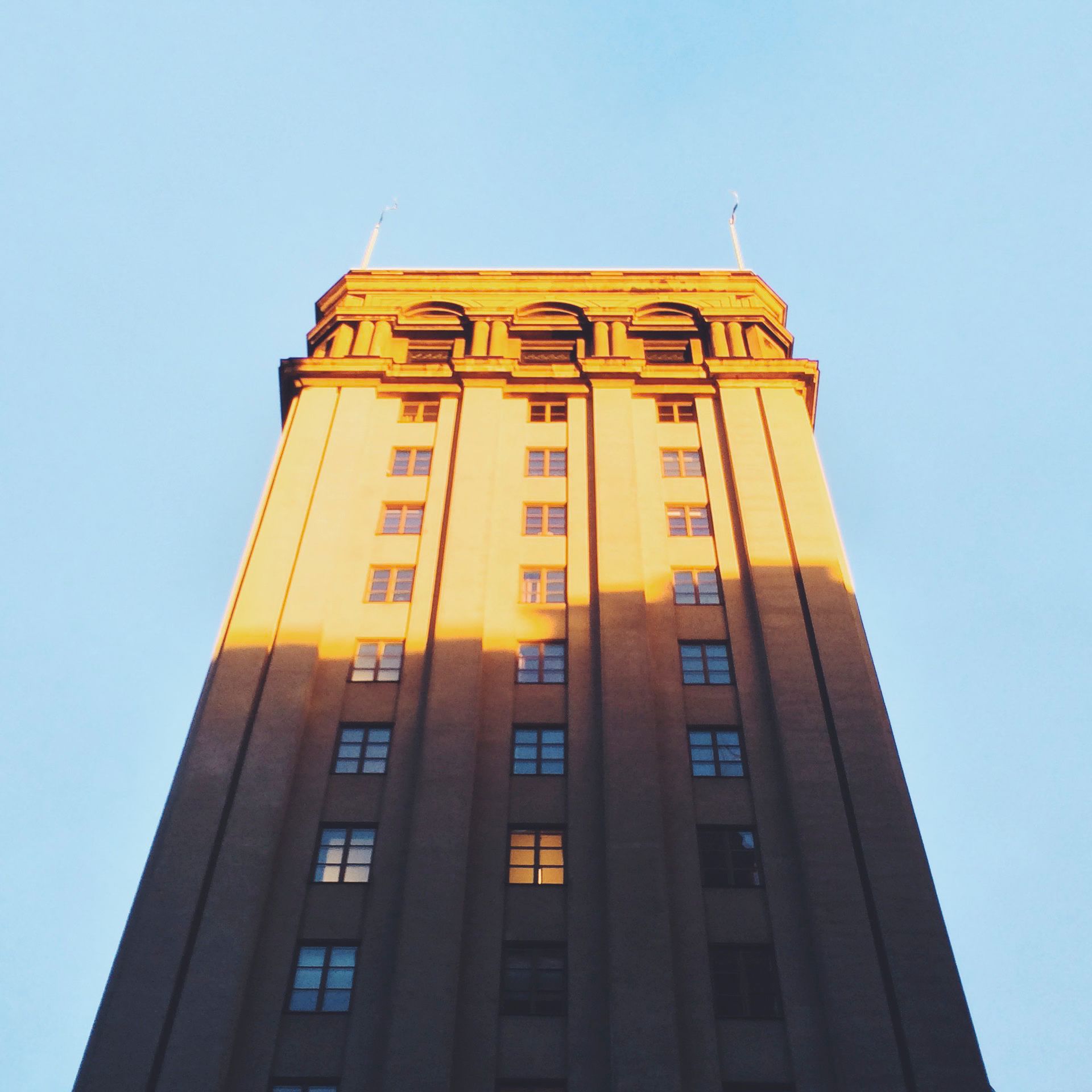 Looking up at a tall stone tower block, its topmost floors lit gold by low sun while the rest of the facade stands in shadow against a pale sky.