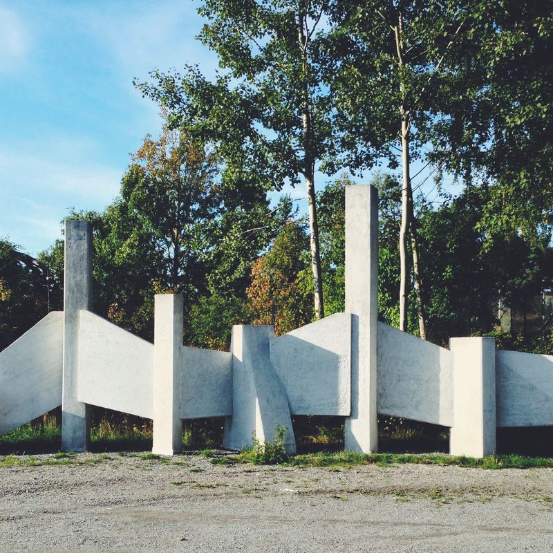 A row of angled white concrete slabs — an outdoor sculpture — standing on gravel in front of birch trees under a blue sky.