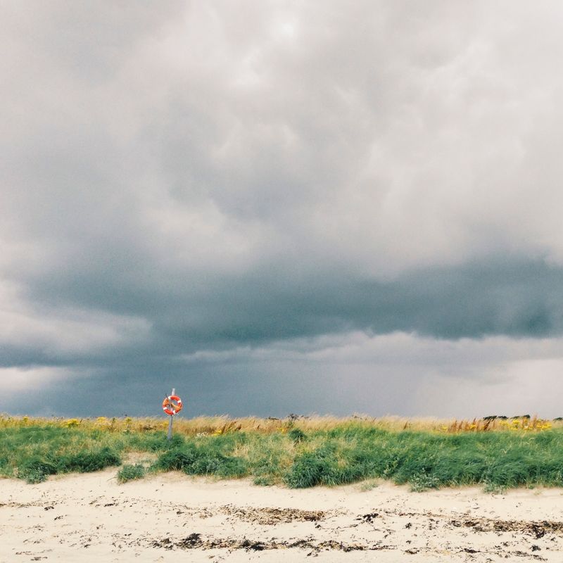 A heavy bank of grey cloud over a beach, with a single orange lifebuoy on a post above the marram grass.