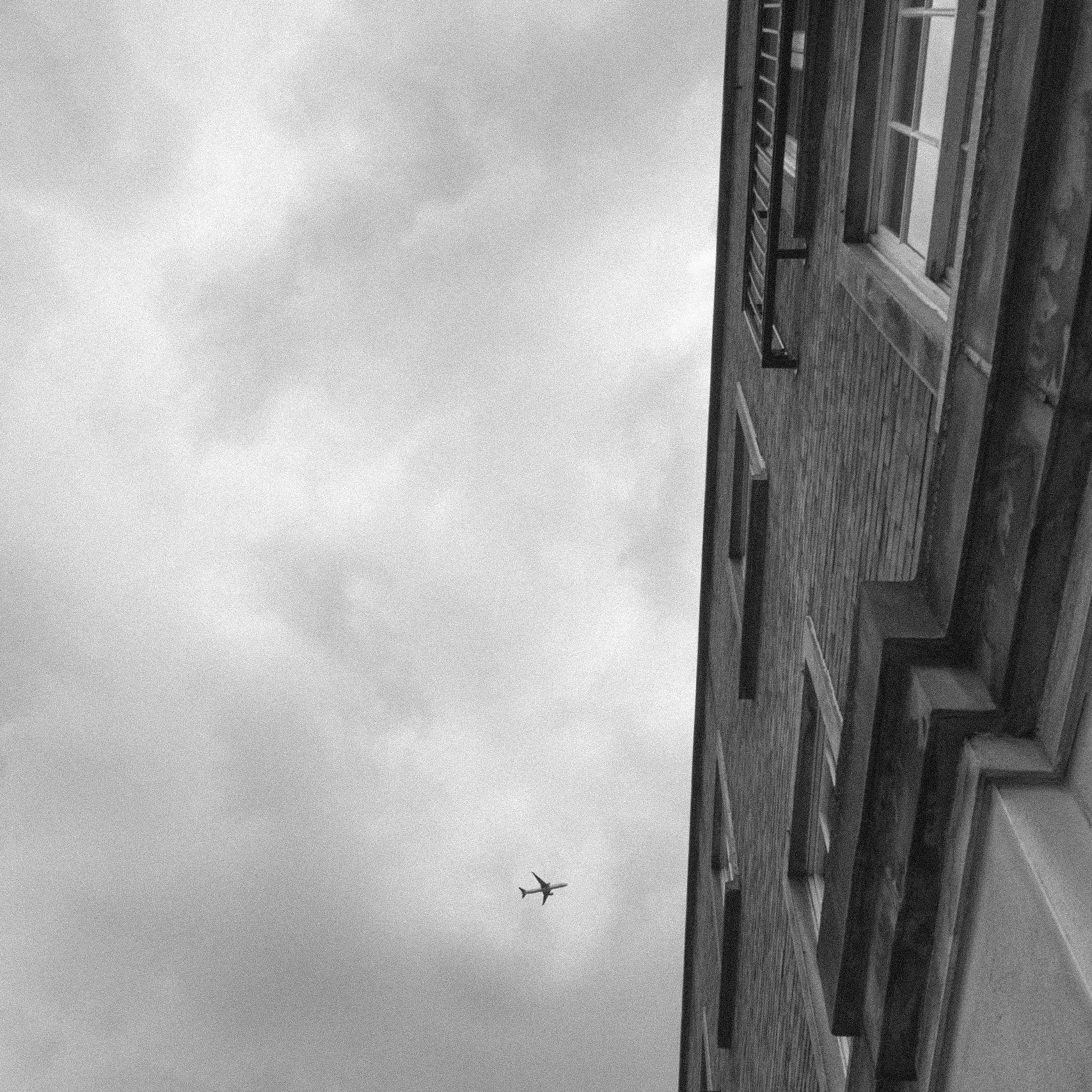 Black and white: a steep upward view along a brick facade of shuttered windows, an airliner crossing the cloudy sky.