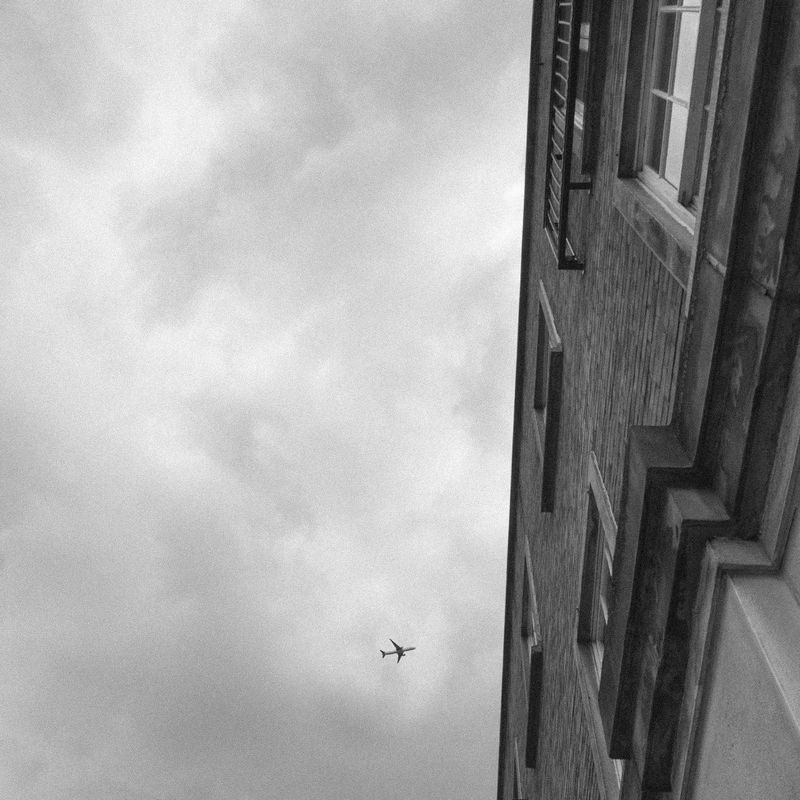 Black and white: a steep upward view along a brick facade of shuttered windows, an airliner crossing the cloudy sky.