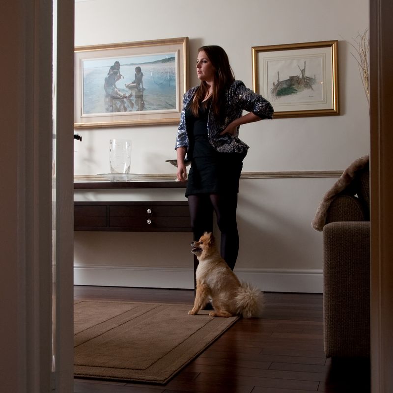 A woman in a sequined jacket stands beside a small dog in a room with two framed pictures and a console table, lit from the left.
