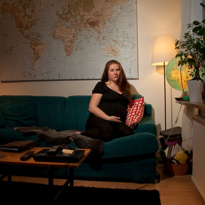 A pregnant woman in a black dress sits on a teal couch in a dimly lit room with a world map on the wall and a floor lamp illuminating a globe.