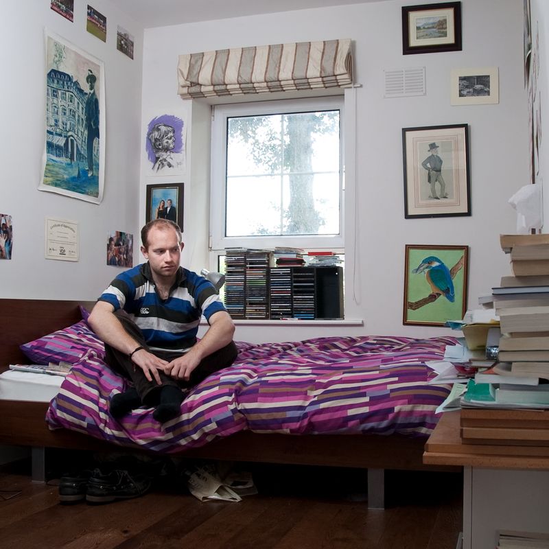 A man in a striped shirt sits on a bed with a purple patterned duvet in a room with a window and various framed pictures and posters on the walls.