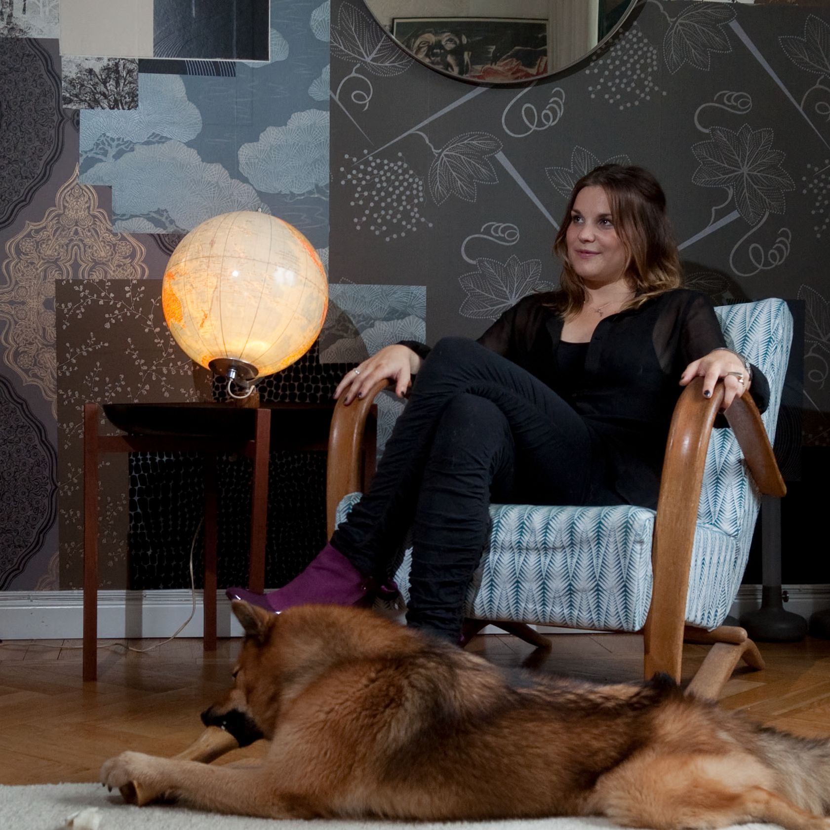 A woman sits in an armchair next to an illuminated globe lamp, with a dog lying on the floor in front of her, against a patterned wallpaper background.