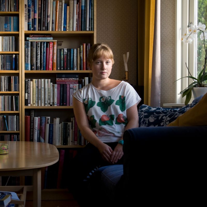 A young woman in a strawberry-patterned shirt sits in a sunlit room with bookshelves and a window.