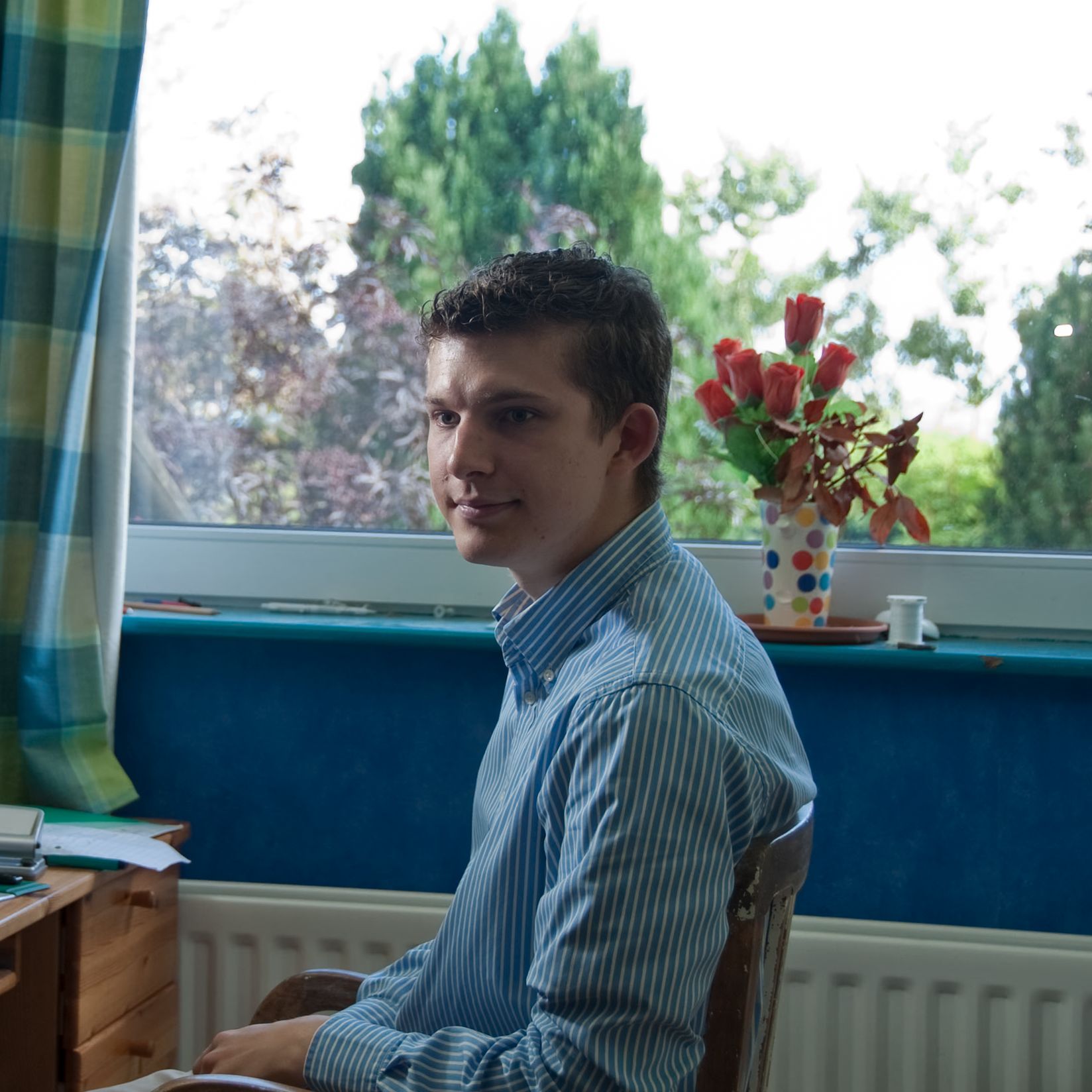 A young man in a striped shirt sits in a wooden chair, looking left, with a window and trees behind him.