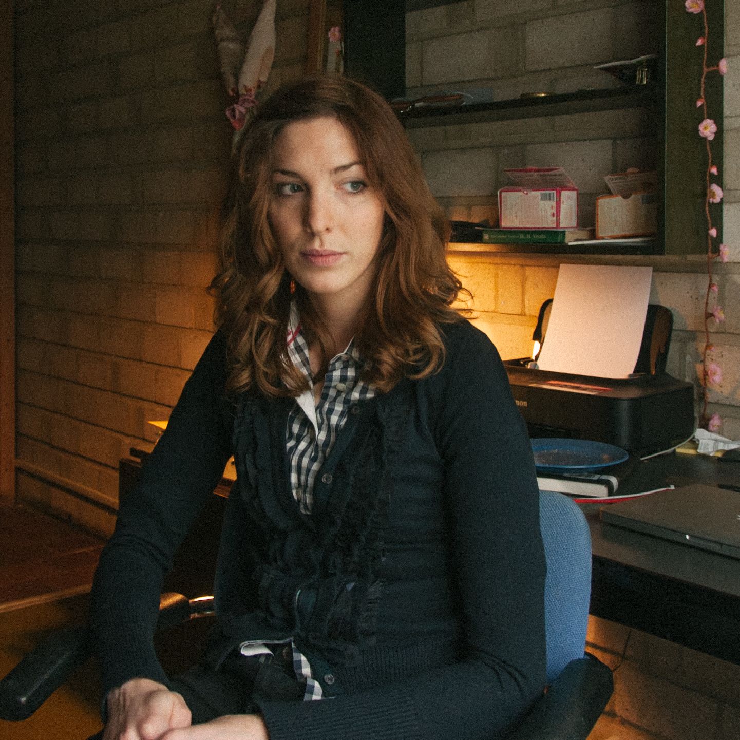 A woman with brown hair sits in a chair, looking right, with a printer and shelves behind her, illuminated by warm light from the left.