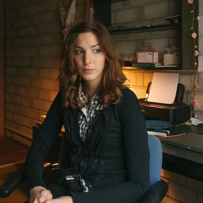 A woman with brown hair sits in a chair, looking right, with a printer and shelves behind her, illuminated by warm light from the left.