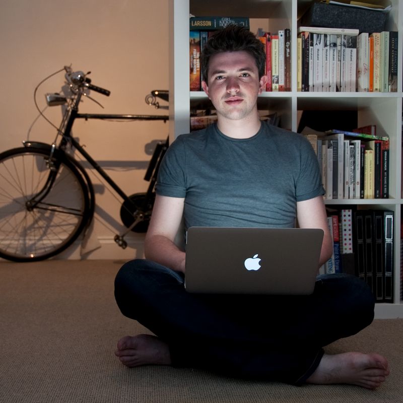 A man sits cross-legged on a carpet with a laptop, a bicycle to his left, and a bookshelf with visible text 